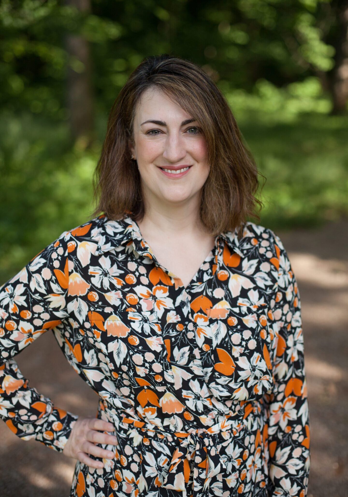 Cassie Headshot A woman in floral shirt standing on dirt path.